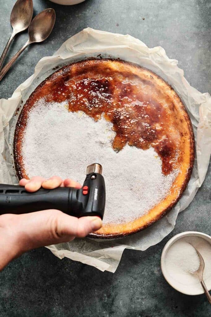 A hand uses a kitchen torch to caramelize sugar on top of a Cr&egrave;me Br&ucirc;l&eacute;e Cheesecake, which sits on parchment paper with a bowl of sugar and two spoons nearby.