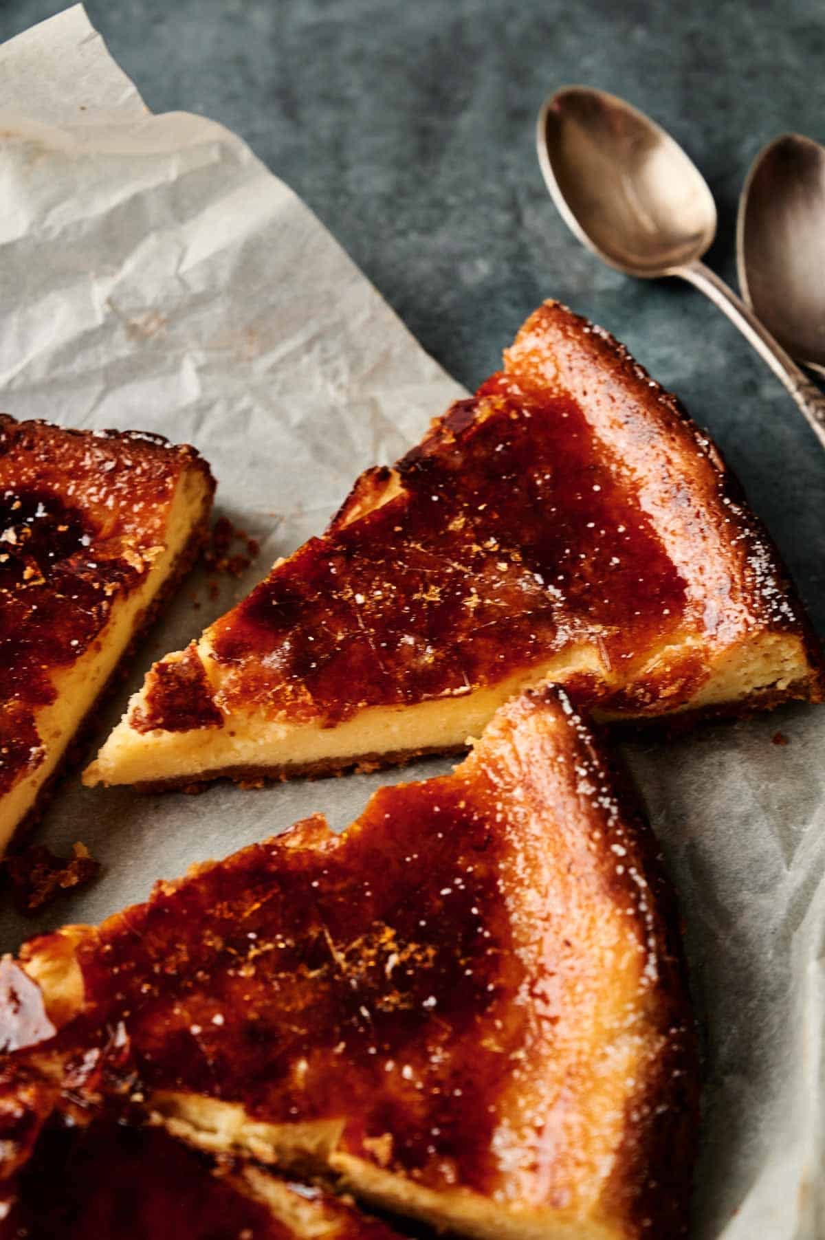 A sliced Cr&egrave;me Br&ucirc;l&eacute;e Cheesecake sits on parchment paper next to two metal spoons on a gray surface.