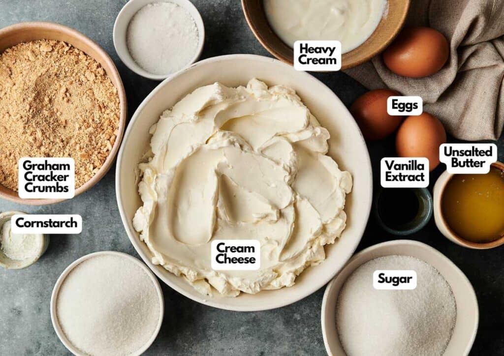 Top-down view of Cr&egrave;me Br&ucirc;l&eacute;e Cheesecake ingredients including cream cheese, graham cracker crumbs, sugar, eggs, unsalted butter, cornstarch, heavy cream, and vanilla extract, each labeled.