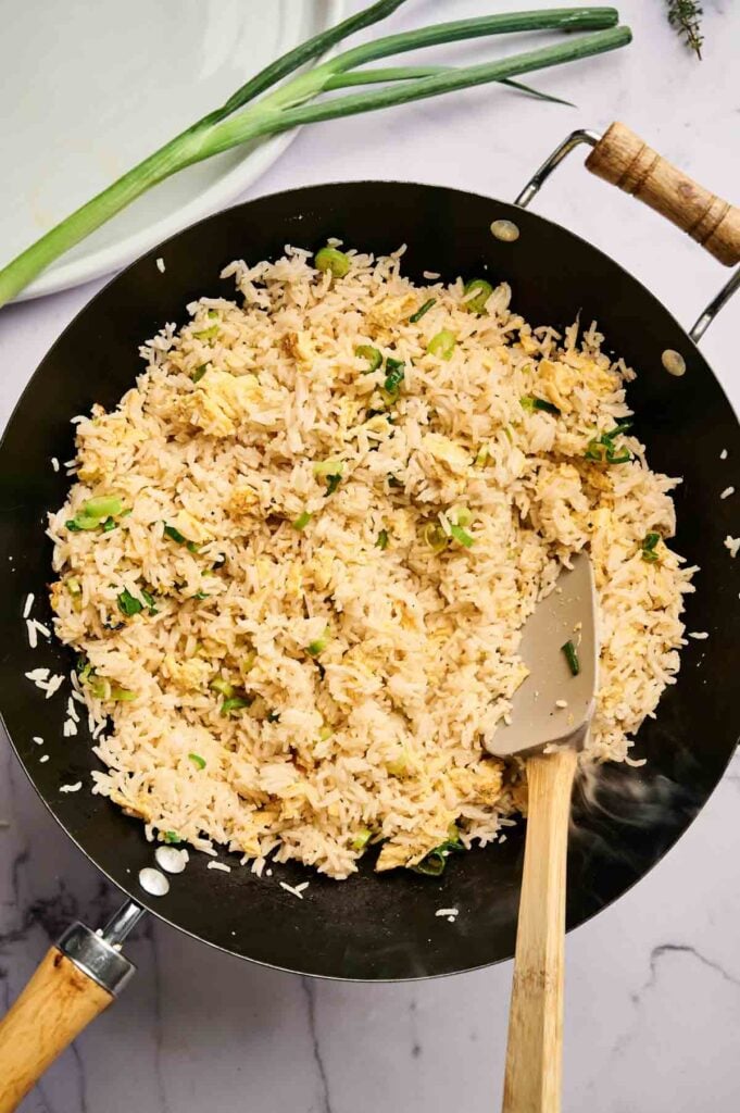 A wok filled with cooked Egg Fried Rice and chopped green onions, with a spatula resting inside; a stalk of green onion lies nearby on a marble surface.