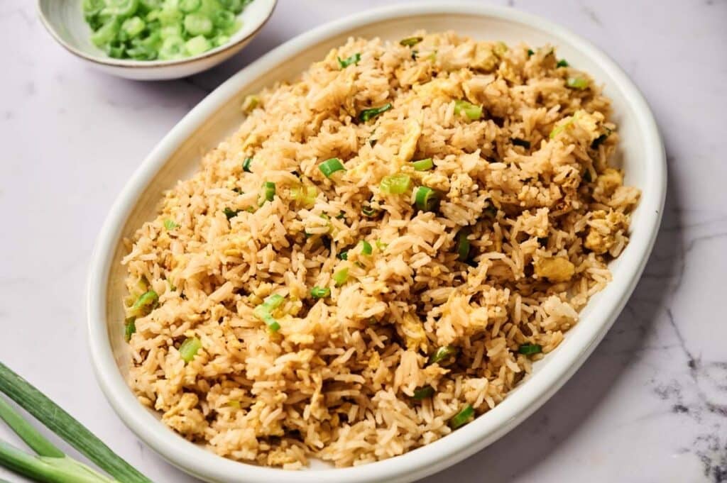 Oval white dish filled with Egg Fried Rice garnished with chopped green onions, placed on a marble surface with a small bowl of green sauce in the background.