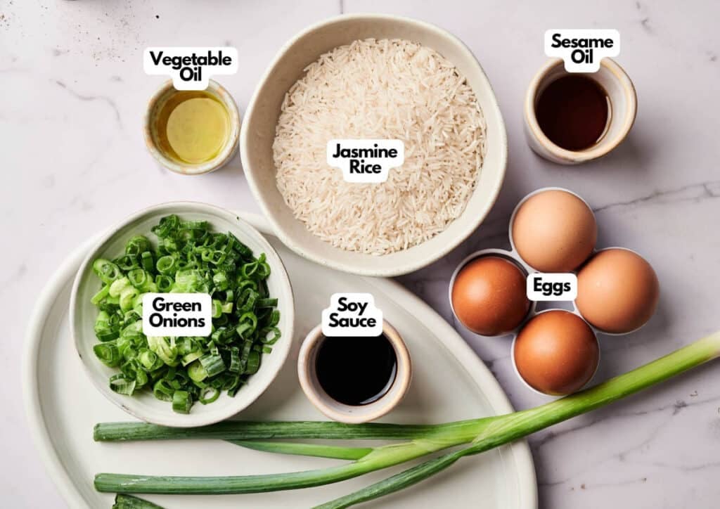 Top-down view of ingredients for Egg Fried Rice in bowls and plates: jasmine rice, eggs, green onions, soy sauce, vegetable oil, and sesame oil on a white surface. Labels clearly identify each item.