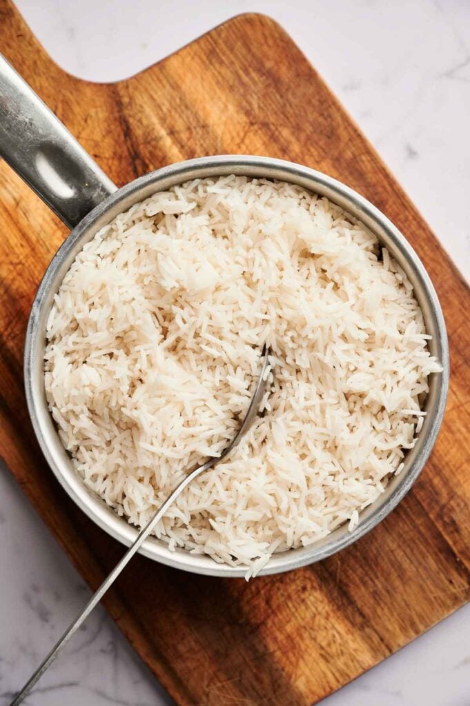 A pot of cooked white rice with a spoon, ready to be transformed into delicious egg fried rice, placed on a wooden cutting board on a marble surface.