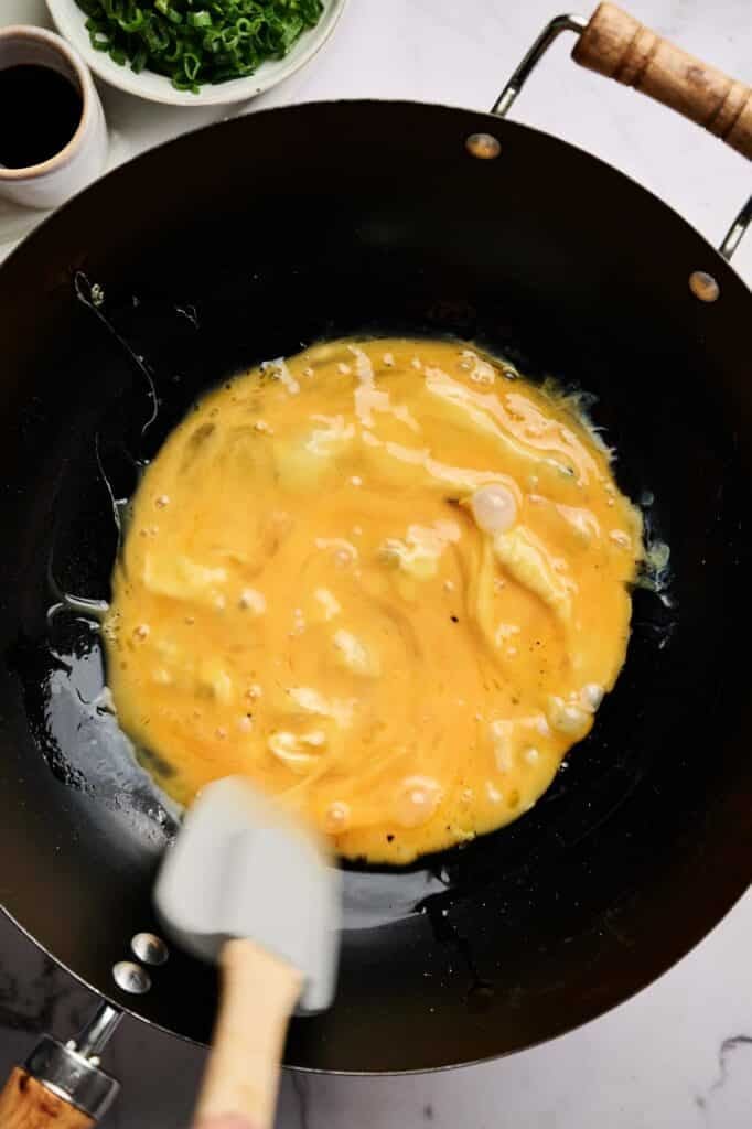 Scrambled eggs being cooked in a black wok, stirred with a spatula—the perfect start for Egg Fried Rice. A bowl of chopped green onions and a cup of liquid are nearby on a white surface.