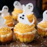 Mini cupcakes with orange and white frosting are topped with white ghost-shaped decorations and white sprinkles, perfect for your Halloween Candy Corn Mini Cheesecakes party display.