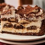 A close-up of a slice of tiramisu cheesecake on a plate, showing layers of coffee-soaked ladyfingers, creamy mascarpone, and a dusting of cocoa powder on top.