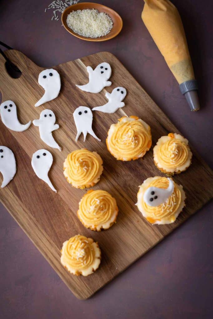 Wooden board with five frosted cupcakes and several ghost-shaped white fondant pieces, next to a piping bag and a small dish of rice—perfect for serving alongside Halloween Candy Corn Mini Cheesecakes.