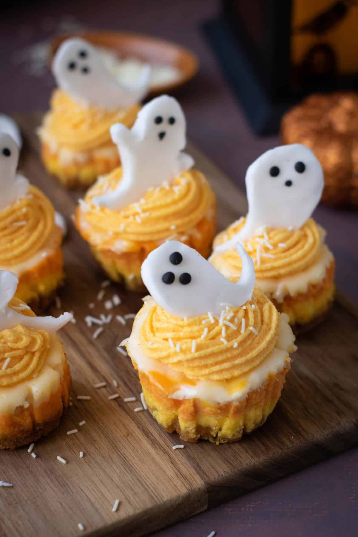 Mini cupcakes with orange frosting and white ghost-shaped toppers, decorated with black eyes, evoke the playful spirit of Halloween Candy Corn Mini Cheesecakes, all arranged on a wooden board with white sprinkles scattered around.
