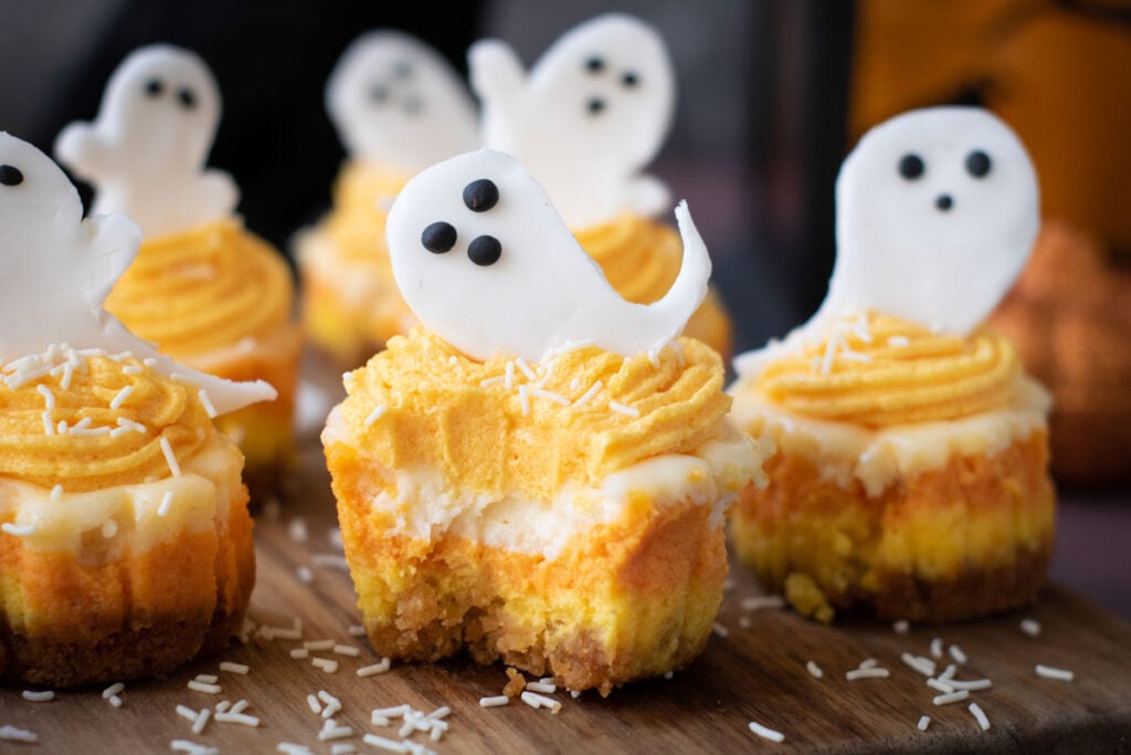 Halloween Candy Corn Mini Cheesecakes feature orange and yellow layers, topped with white icing ghosts and sprinkles, all displayed on a wooden board.