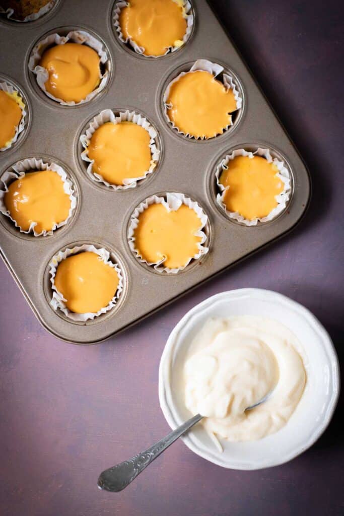 A muffin tin filled with cupcake liners for Halloween Candy Corn Mini Cheesecakes sits next to a white bowl of creamy mixture with a spoon on a purple surface.