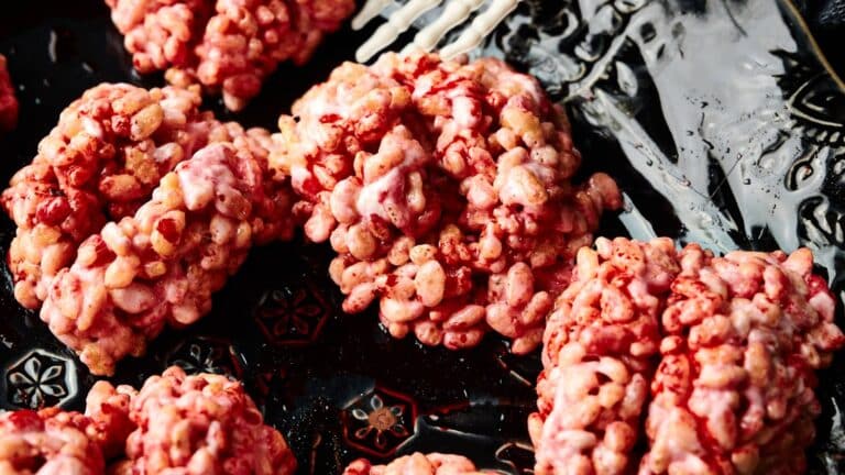 Close-up of several pinkish-red rice cereal treats on a dark, patterned plate with a white plastic skeleton hand.