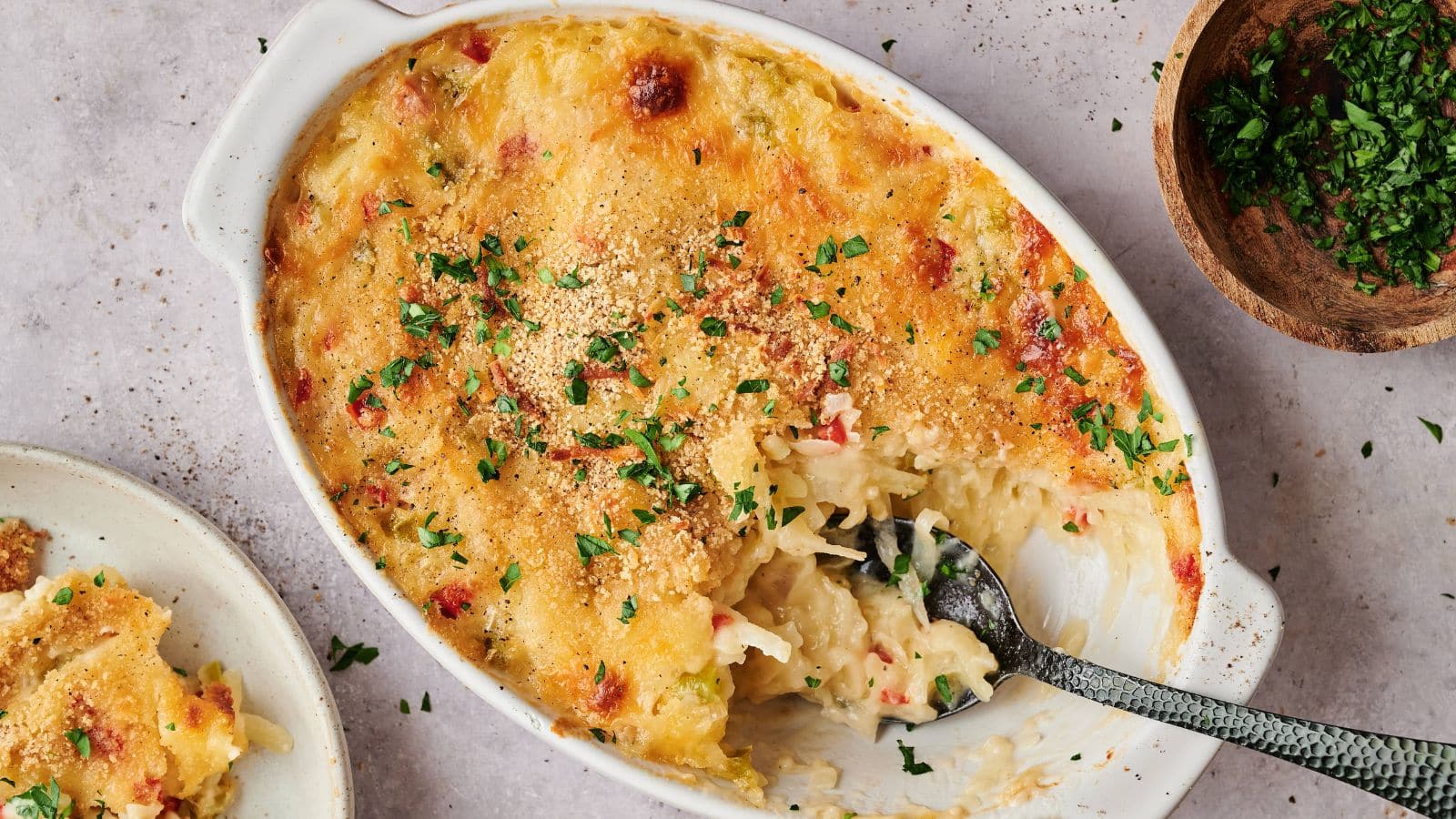 A baked casserole dish topped with melted cheese and chopped herbs, with a serving spoon inside and a portion removed; a bowl of herbs is nearby.