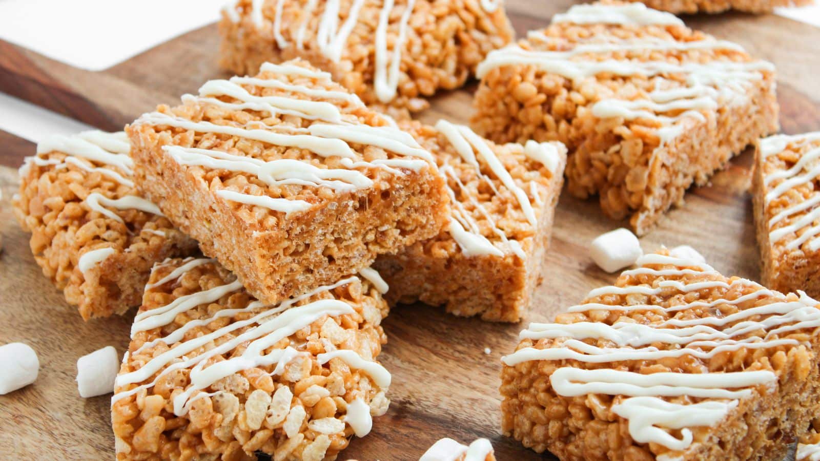 Rectangular rice crispy treats drizzled with white icing are arranged on a wooden surface, with small marshmallows scattered nearby.