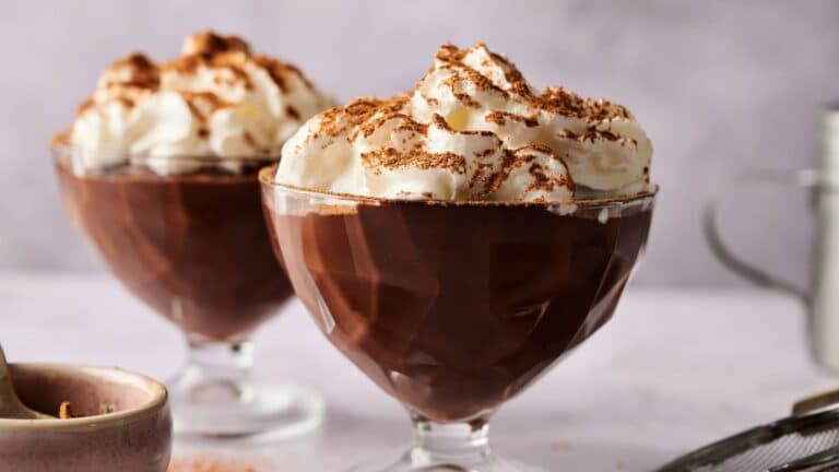 Two glass dessert cups filled with chocolate pudding, topped with whipped cream and a sprinkle of cocoa powder, sit on a light surface.