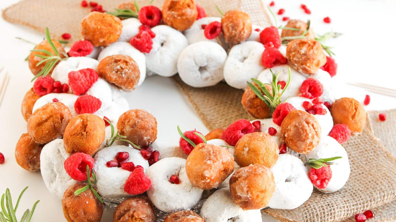A wreath made of powdered and glazed donut holes, garnished with fresh raspberries, pomegranate seeds, and rosemary sprigs, displayed on a burlap ribbon.