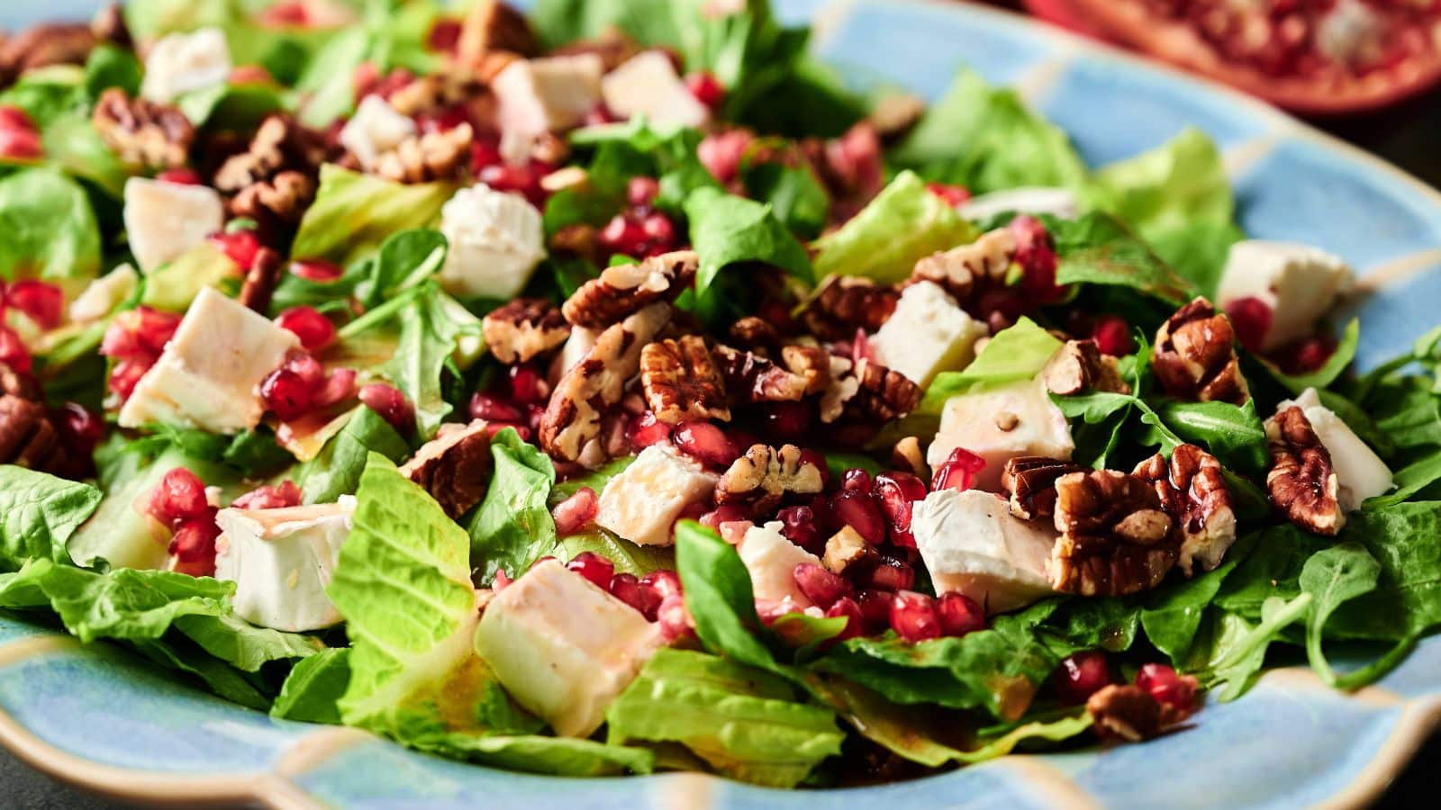 A close-up of a salad with chopped romaine lettuce, diced white cheese, pecans, and pomegranate seeds on a blue plate.