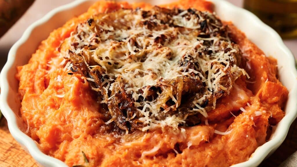 A dish of mashed sweet potatoes topped with shredded cheese and caramelized onions in a white bowl.