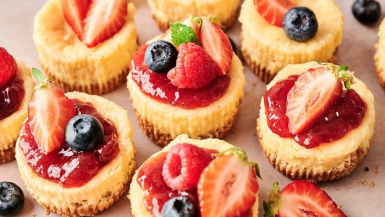 Mini cheesecakes topped with strawberry sauce and fresh berries, including strawberries, raspberries, and blueberries, arranged on a light-colored surface.