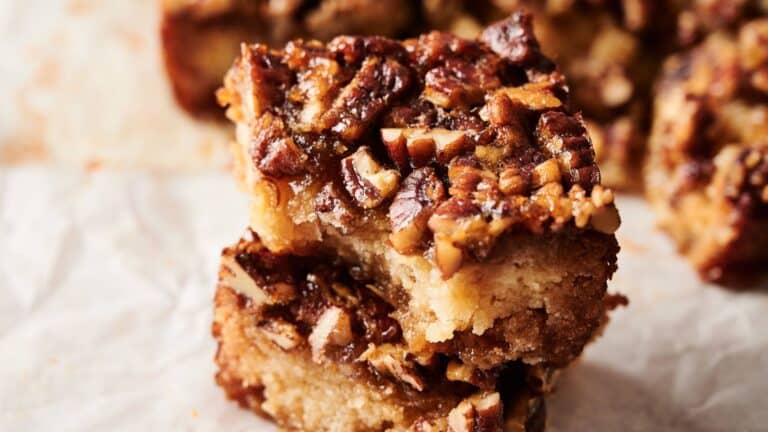 Two pecan-topped dessert bars are stacked on parchment paper, showing a dense, crumbly base and a caramelized nut topping.