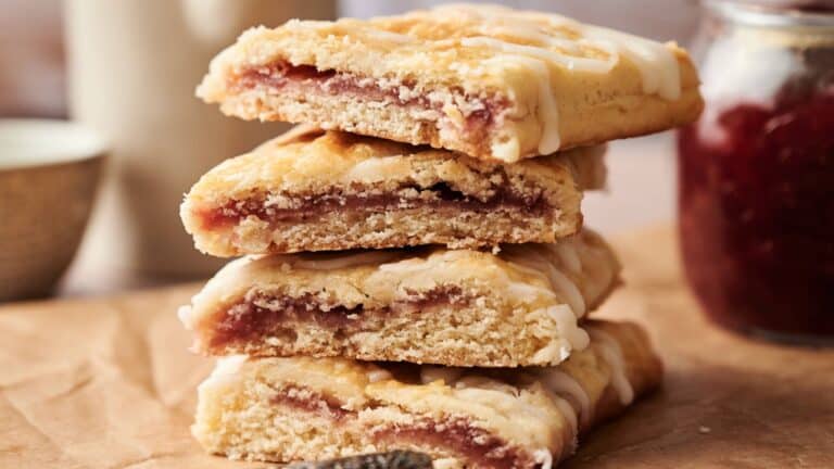 A stack of rectangular pastries with a crumbly crust, filled with a red fruit jam and drizzled with white icing, placed on brown parchment paper.