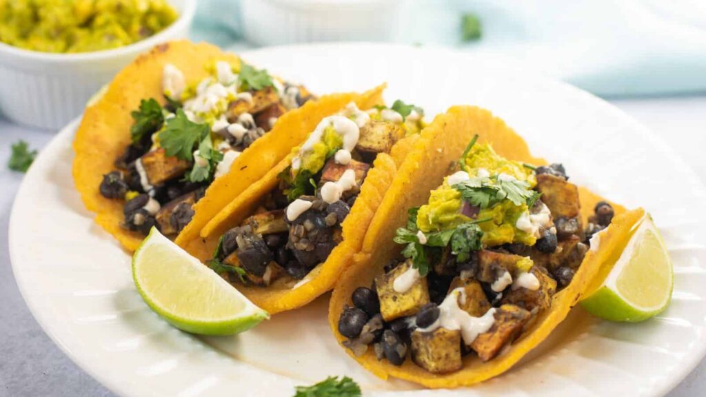 Three tacos filled with black beans, tofu, and vegetables, topped with cilantro, sauce, and guacamole, served on a white plate with lime wedges.