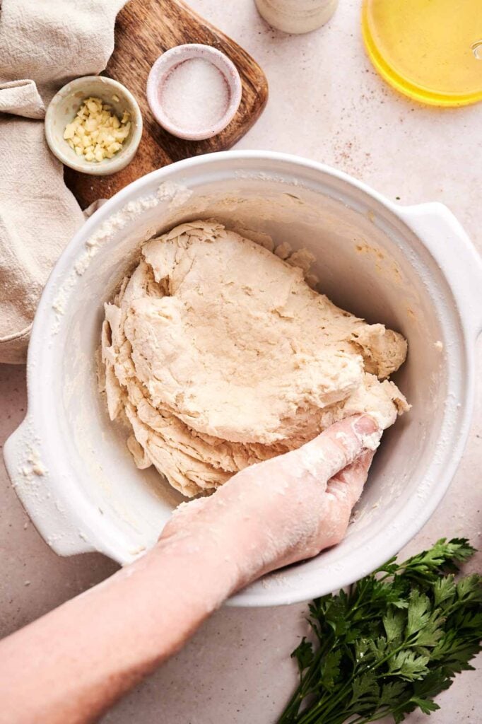 A hand kneads naan bread dough in a white bowl on a countertop, surrounded by parsley, chopped garlic, salt, oil, and a wooden board.