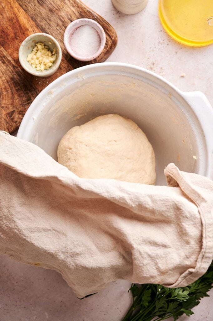 A bowl with naan bread dough covered partially by a cloth sits on a countertop next to chopped garlic, salt, a wooden board, and fresh herbs.