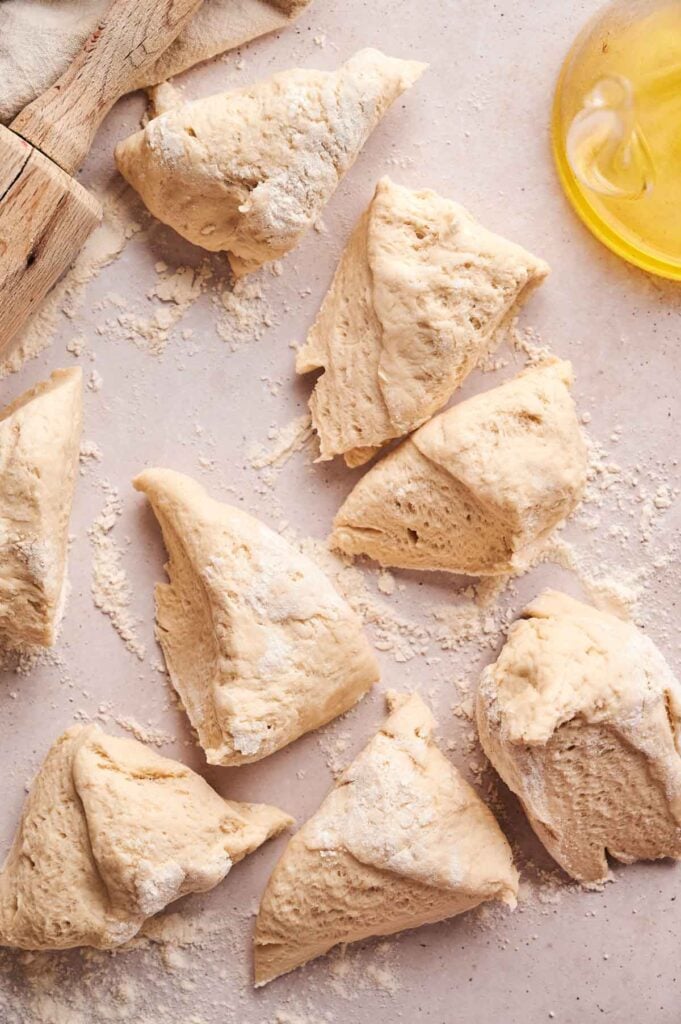 Several pieces of raw naan bread dough are cut and scattered on a floured surface, with a small dish of olive oil and a wooden utensil visible nearby.