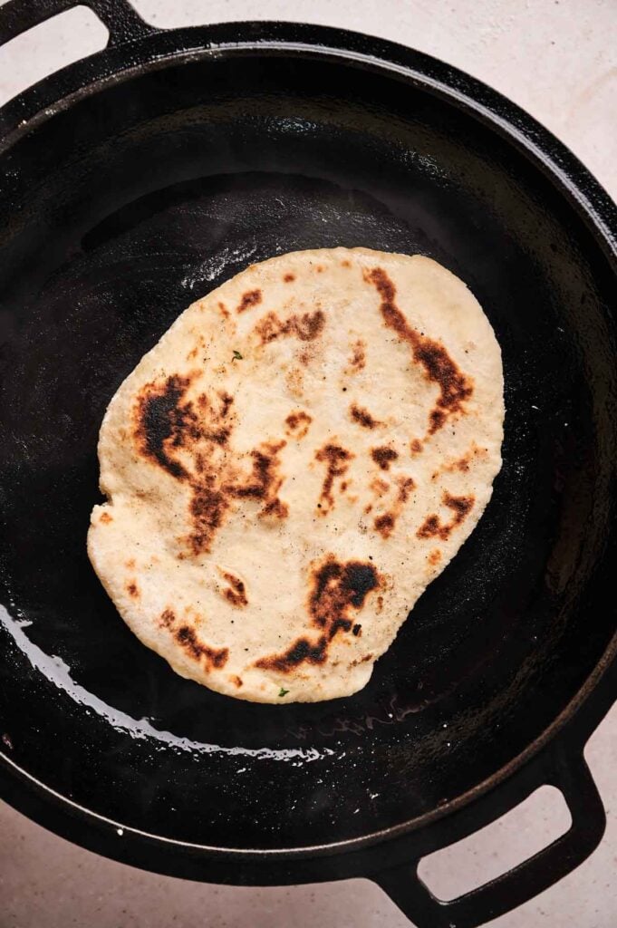 Naan bread cooking in a black cast iron skillet, showing enticing areas of browning and slight charring on the surface.
