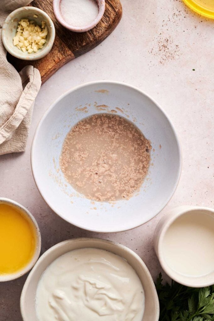 A bowl with a yeast mixture for Naan Bread sits on a counter, surrounded by small bowls of melted butter, yogurt, milk, chopped garlic, and a dish of salt.