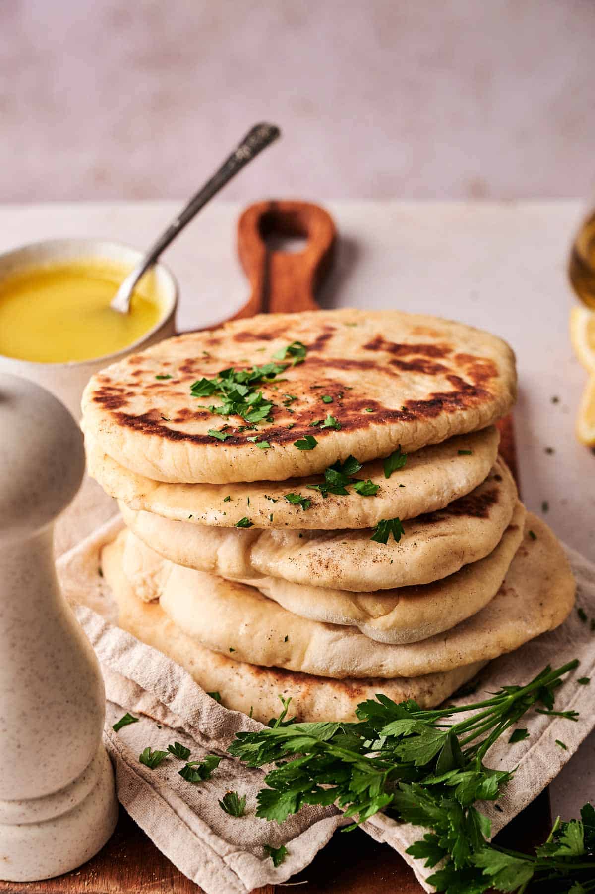 A stack of naan bread garnished with chopped parsley sits on a cloth, with fresh herbs, a bowl of melted butter, and a pepper grinder nearby.