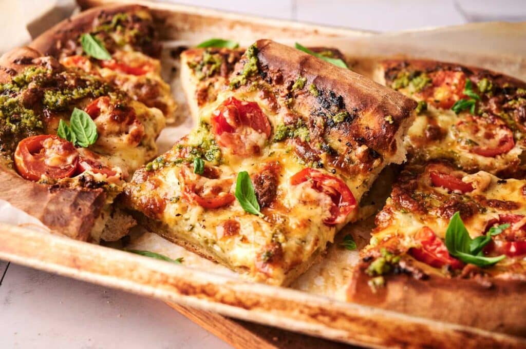 Rectangular slices of homemade pesto pizza topped with melted cheese, cherry tomatoes, and fresh basil leaves on a parchment-lined baking sheet.