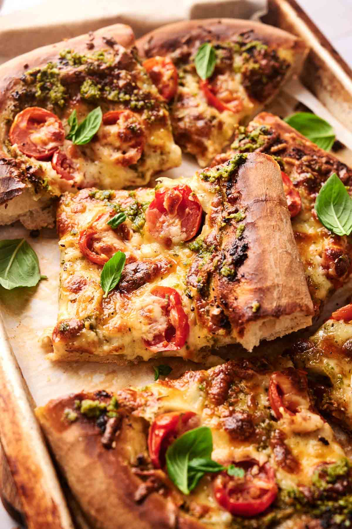 Rectangular slices of homemade pesto pizza with a thick crust, topped with melted cheese, cherry tomatoes, pesto, and fresh basil leaves on a baking tray.