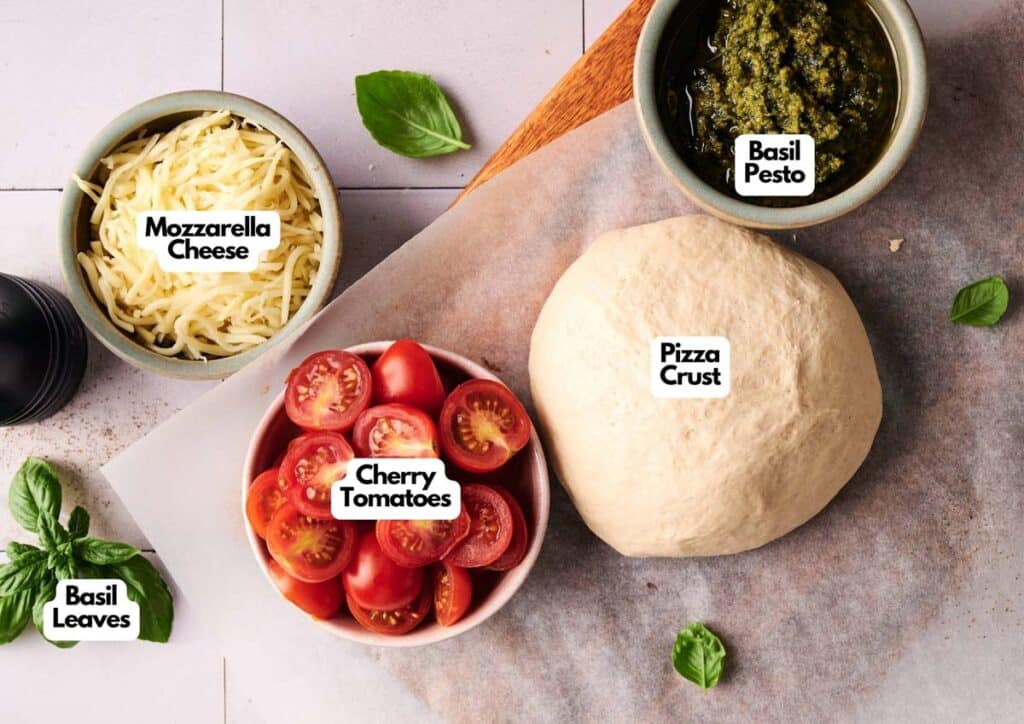 A flat lay of ingredients for Pesto Pizza: shredded mozzarella cheese, basil pesto, pizza crust dough, sliced cherry tomatoes, and fresh basil leaves.
