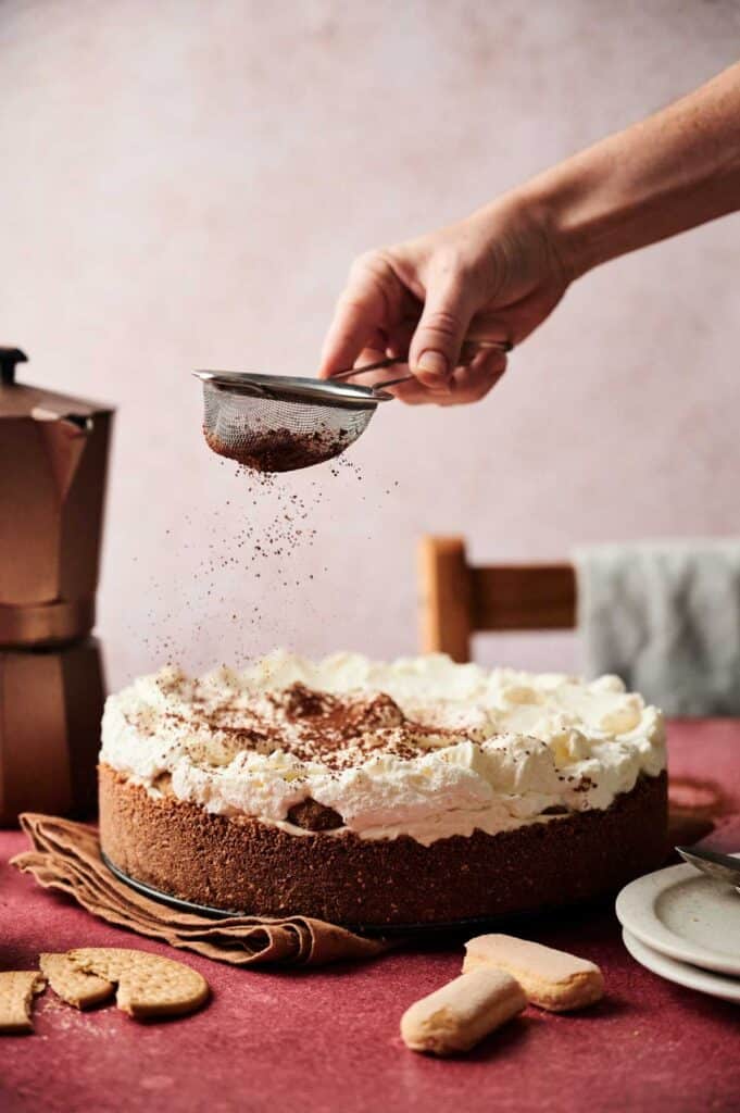 A hand sprinkles cocoa powder over a whipped cream-topped Tiramisu Cheesecake on a table, with a coffee pot, napkin, and cookies nearby.