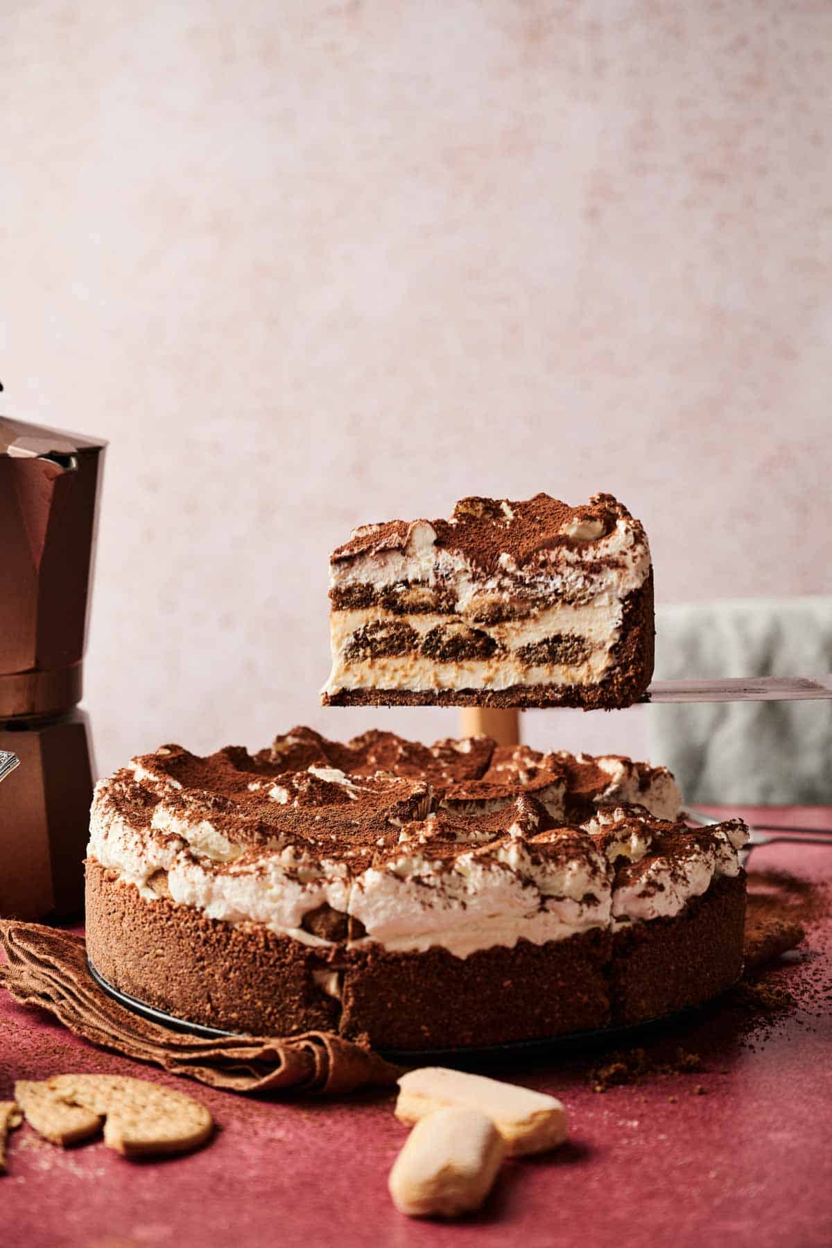 A slice of layered tiramisu cheesecake is being lifted from the whole cake, which sits on a table with some cookies and a coffee pot nearby.