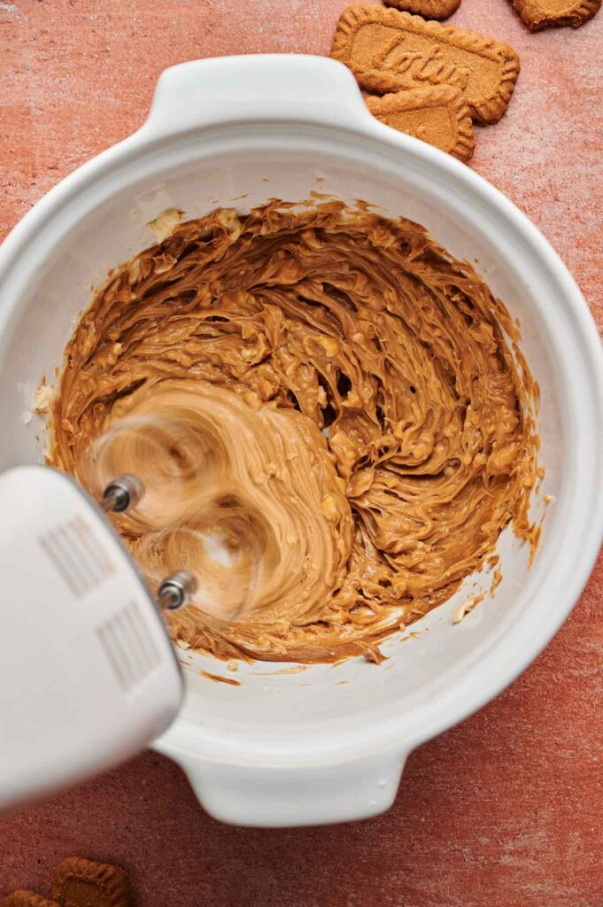 An electric hand mixer blends a thick, brown mixture in a white bowl, with Biscoff Sandwich Cookies nearby on a reddish surface.