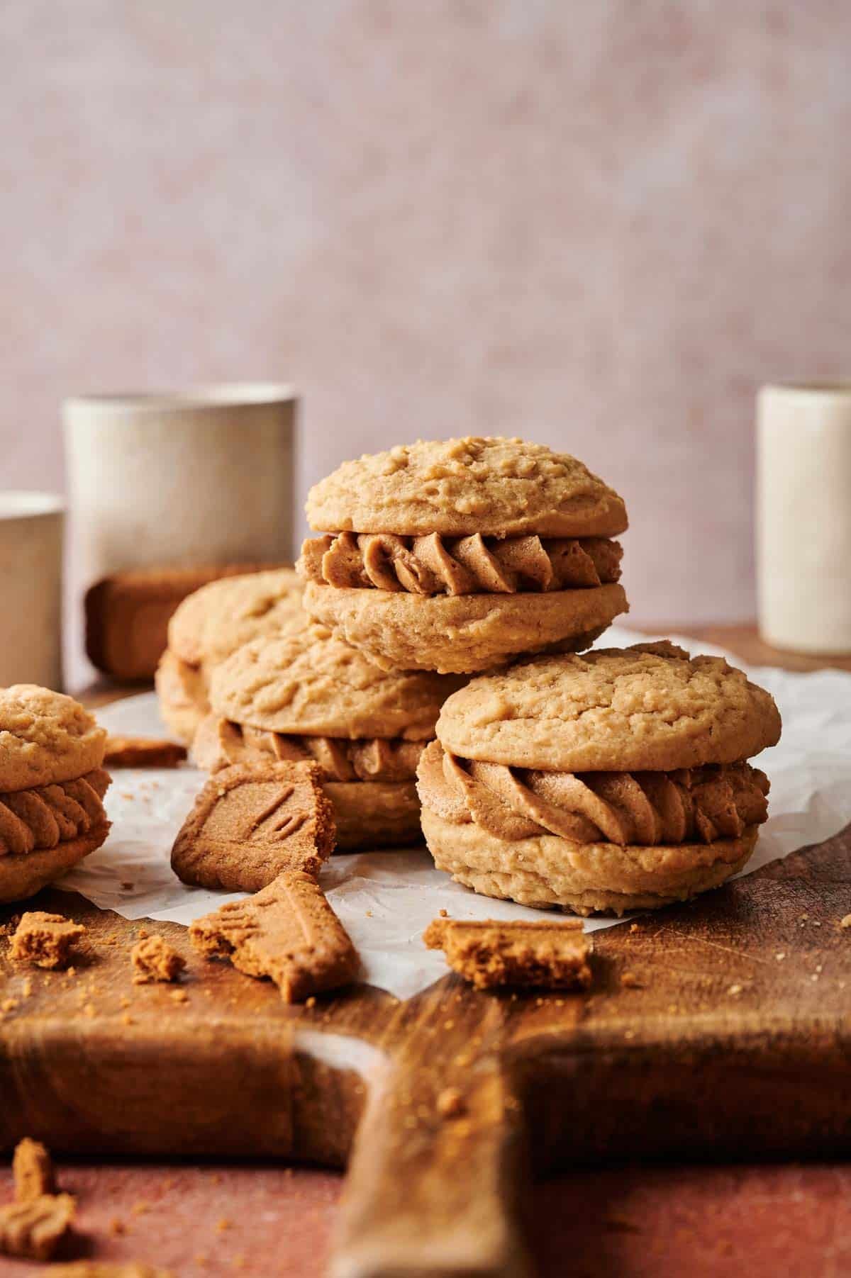 A stack of Biscoff Sandwich Cookies with a creamy filling sits on a wooden board, surrounded by cookie crumbs and mugs in the background.