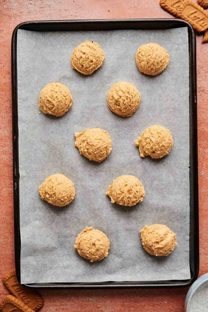 A baking tray lined with parchment paper holds eleven evenly spaced scoops of unbaked Biscoff Sandwich Cookie dough.