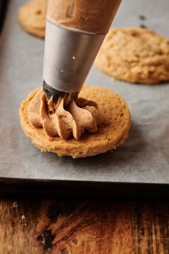 A piping bag is applying frosting onto a round Biscoff Sandwich Cookie on a parchment-lined baking sheet.