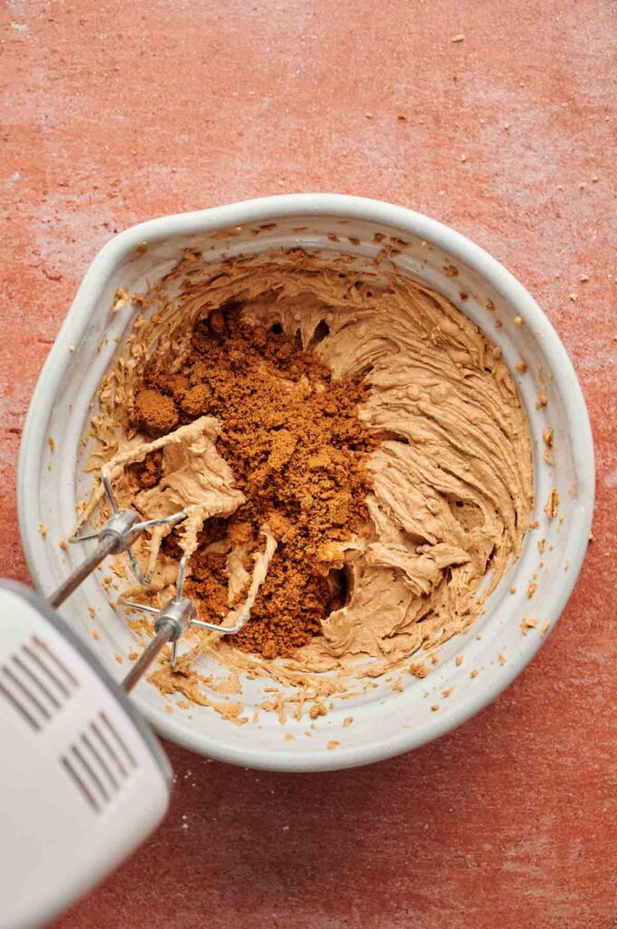 An electric hand mixer blends Biscoff Sandwich Cookies with other brown ingredients in a white mixing bowl on a reddish countertop.