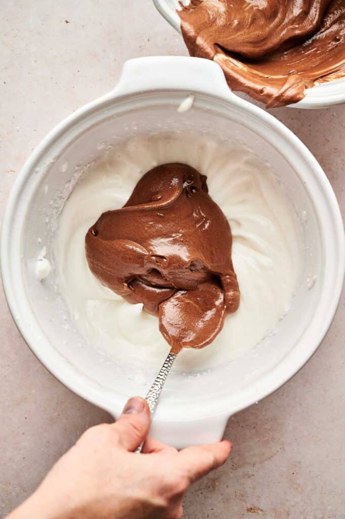 A person's hand gently folds rich chocolate mousse into whipped egg whites in a white mixing bowl using a spoon.