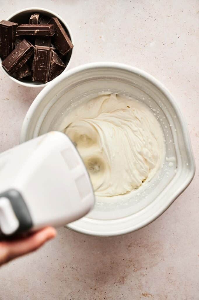 A hand mixer is whipping cream in a white bowl, preparing the base for chocolate mousse, with a small bowl of chocolate pieces nearby on a light countertop.