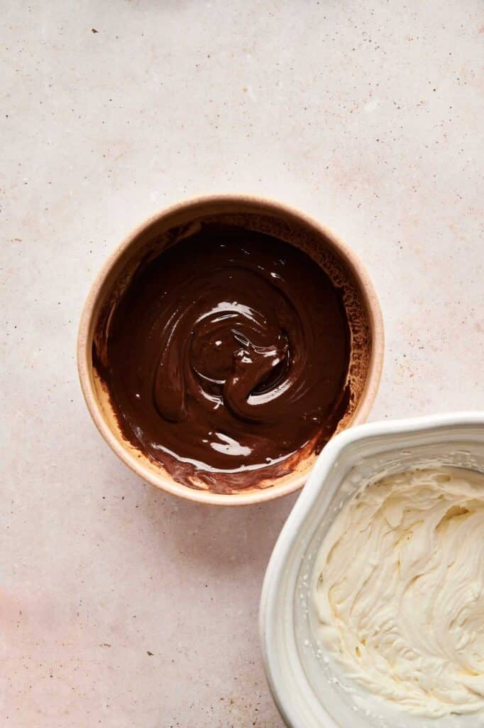 A bowl of melted chocolate sits next to a bowl of whipped cream on a light-colored surface, ready to be combined into a luscious Chocolate Mousse.