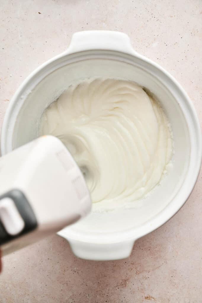 A hand mixer blends a creamy white mixture for Chocolate Mousse in a large white mixing bowl on a light-colored surface.