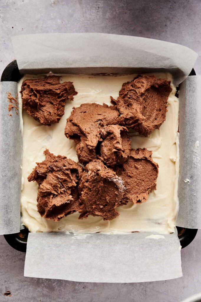 A baking pan lined with parchment paper holds a layer of cream-colored batter for Cream Cheese Brownies, topped with several dollops of chocolate batter, ready to be spread.