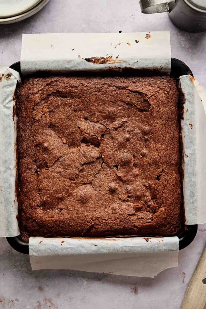A square batch of cream cheese brownies in a parchment-lined baking pan, freshly baked with a beautifully cracked top surface.