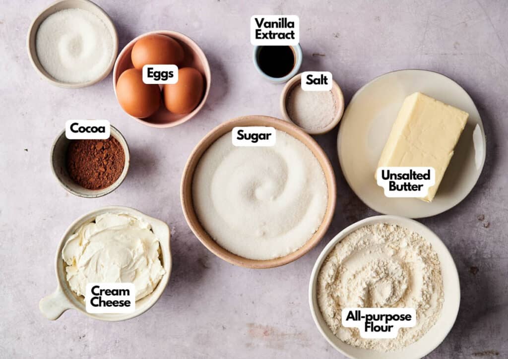 Assorted baking ingredients for Cream Cheese Brownies are labeled: eggs, vanilla extract, salt, unsalted butter, all-purpose flour, sugar, cocoa, and cream cheese&mdash;artfully arranged on a light surface.