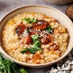 A bowl of creamy Butternut Squash Risotto is topped with roasted squash, grated cheese, and fresh parsley, with a cloth napkin and herbs nearby.