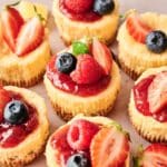Mini Cheesecakes topped with strawberry sauce, halved strawberries, raspberries, and blueberries are arranged closely on a light surface.